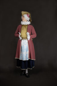 fotografia rekonstrukcji ubioru kobiety żydowskiej z przełomu XIX i XIX wieku, Na czarnym tle sylwetka kobiety w czerwonym długim kaftanie, granatowej spódnicy, białym wzorzystym fartuchu i czepcu ze złotymi dodatkami. 