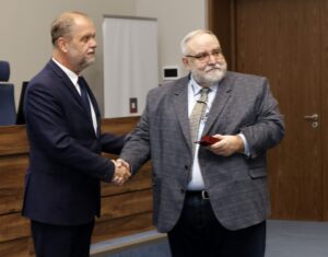 Dyrektor Archiwum Narodowego w Krakowie prof. Wojciech Krawczuk wręcza Romanowi Marcinkowi odznakę „Za zasługi dla archiwistyki”. Mężczyźni stoją w sali konferencyjnej i podają sobie ręce; jeden z nich trzyma w drugiej ręce małe pudełko z odznaczeniem. W tle widać drewniany stół, krzesła oraz drzwi.