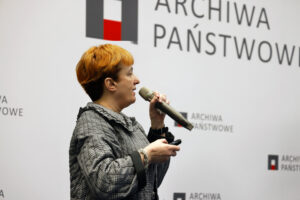Dr Kamila Follprecht, zastępca dyrektora Archiwum Narodowego w Krakowie, podczas wystąpienia „Zbiory archiwów państwowych jako źródło wiedzy zabytkoznawczej na przykładzie Krakowa” w ramach III Kongresu Konserwatorów Polskich. Prelegentka stoi na scenie przy mównicy, przemawia do mikrofonu, a w tle widoczny jest ekran z prezentacją i logo wydarzenia.