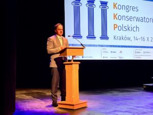 Wystąpienie Naczelnego Dyrektora Archiwów Państwowych dr. Pawła Pietrzyka podczas uroczystego otwarcia obrad III Kongresu Konserwatorów Polskich. Mężczyzna w jasnoszarym garniturze stoi przy drewnianej mównicy na scenie i przemawia do mikrofonu. W tle widać duży ekran z logo wydarzenia i napisem „Kongres Konserwatorów Polskich, Kraków, 14–16 X 2025